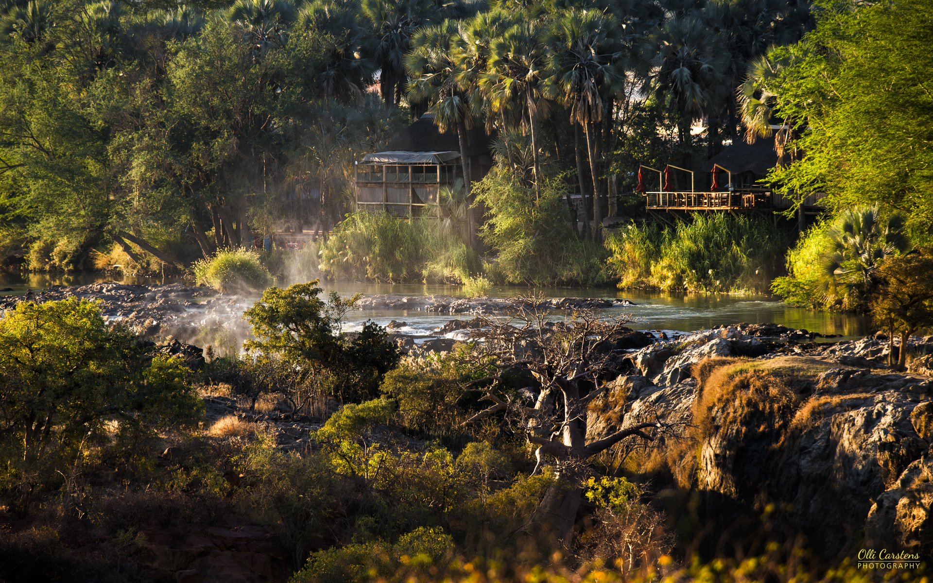 Epupa Falls Ein ruhiger Fluss fließt durch einen Wald mit grünen Bäumen und felsigem Ufer.